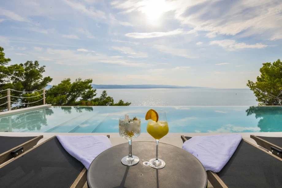 Sun loungers and a table with two cocktails sit by the pool, showcasing a beautiful view of the sea and island
