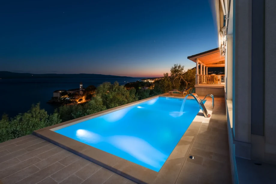 Infinity pool of Villa Bol Summer House with glowing blue water on a terrace, overlooking a serene sea and distant hills at twilight. A cozy, well-lit veranda is visible nearby.