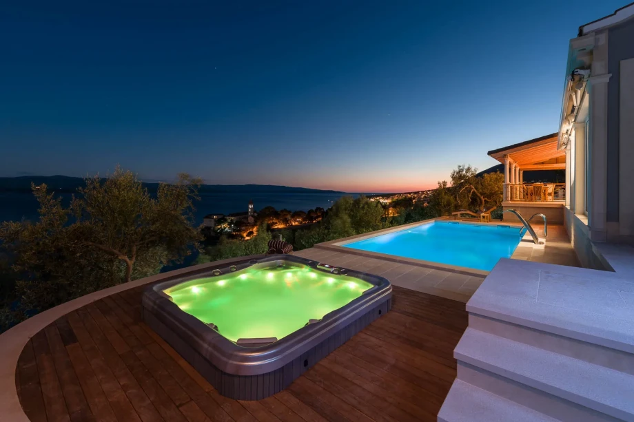 Luxurious family villa terrace at dusk with a glowing jacuzzi and pool. Overlooks serene sea and distant city lights, creating a tranquil, inviting atmosphere.