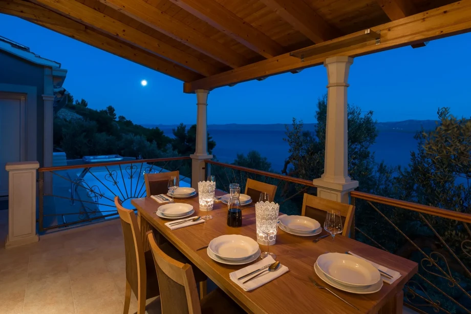Outdoor dining area in pet friendly Villa Bol Summer House overlooking a serene, moonlit sea. A wooden table set with white dishes, glasses, and a carafe exudes a cozy, inviting ambiance.