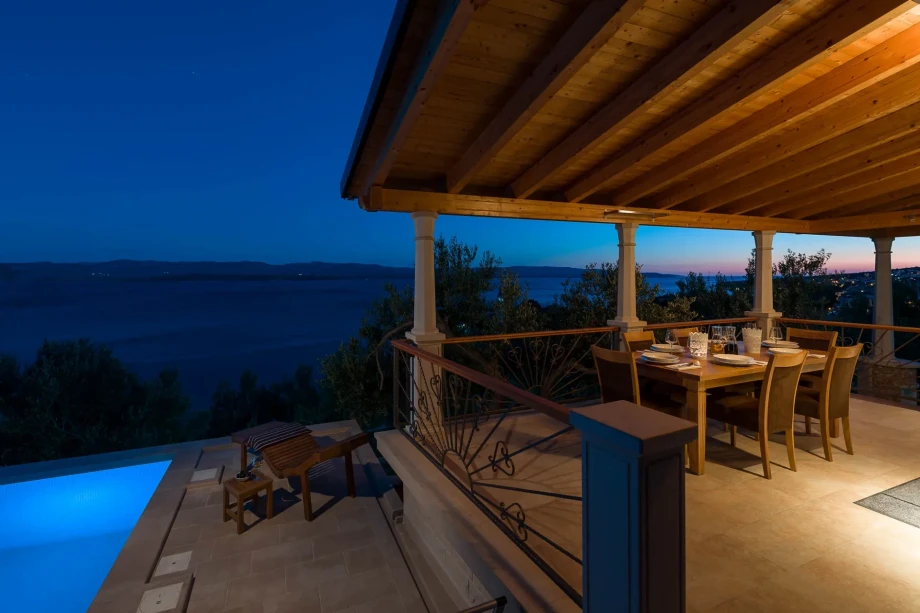 A covered patio with a wooden dining table set for dinner overlooks a serene, illuminated pool and a tranquil ocean view at twilight, evoking a sense of relaxation.