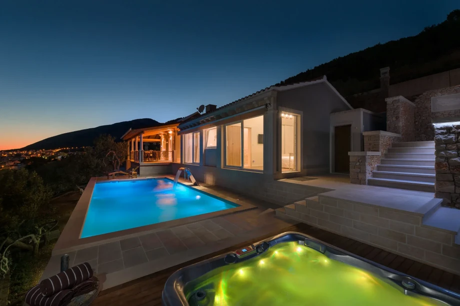 Modern villa at dusk, with an illuminated pool and hot tub on the patio. Warm interior lights glow through windows, with a serene mountain view.
