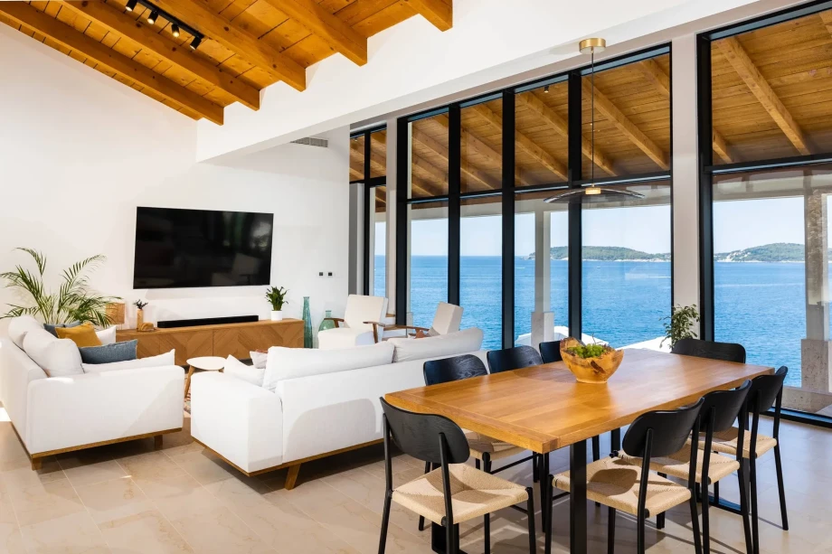A beautiful living room in the luxury villa My Place features a sofa, a wall-mounted TV, a table, chairs, and a stunning open view of the sea.