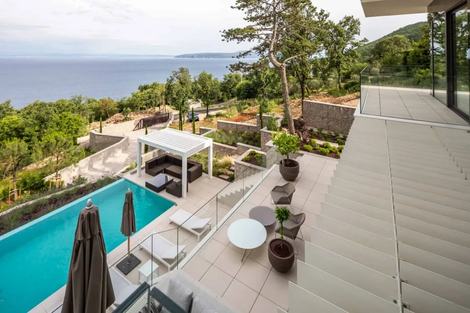 View from the terrace overlooking a serene sea, with a sleek pool, loungers, and a shaded seating area. Lush greenery and hills create a tranquil atmosphere.