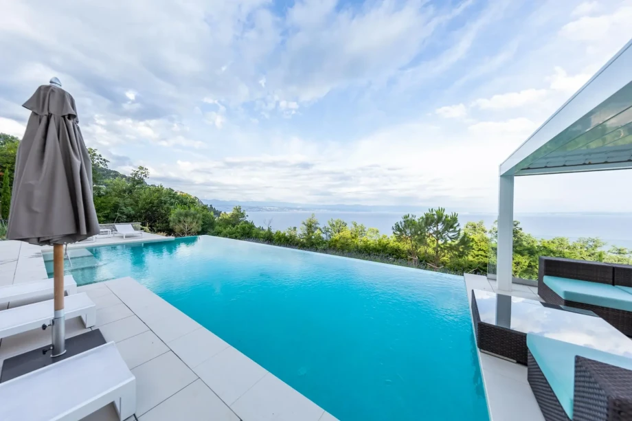 Infinity pool overlooking the sea and lush greenery under a partly cloudy sky. White loungers and a shaded seating area create a serene, luxurious ambiance.