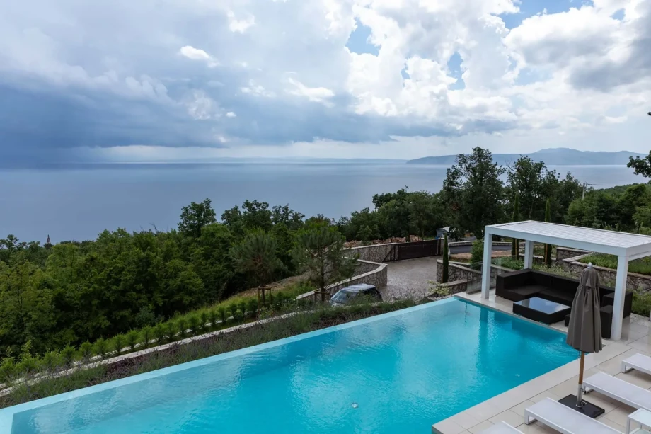 Infinity pool overlooking a stunning sea view, set against a backdrop of cloudy skies, surrounded by lush greenery, and featuring a modern seating area.