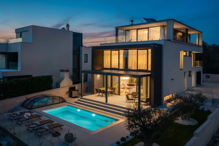 Night view on the property of luxury Villa Happiness in Petrcane with sun loungers, covered terrace with dining table and illuminated swimming pool