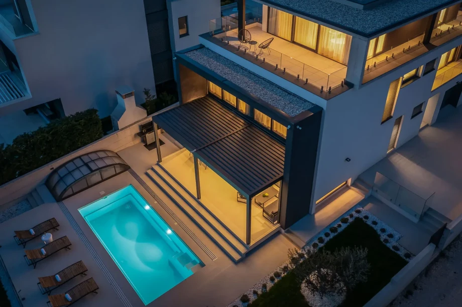 Aerial night view on the outdoor fenced villa area, pool with the patio and sun loungers, main terrace, and bedroom terrace with sitting furniture